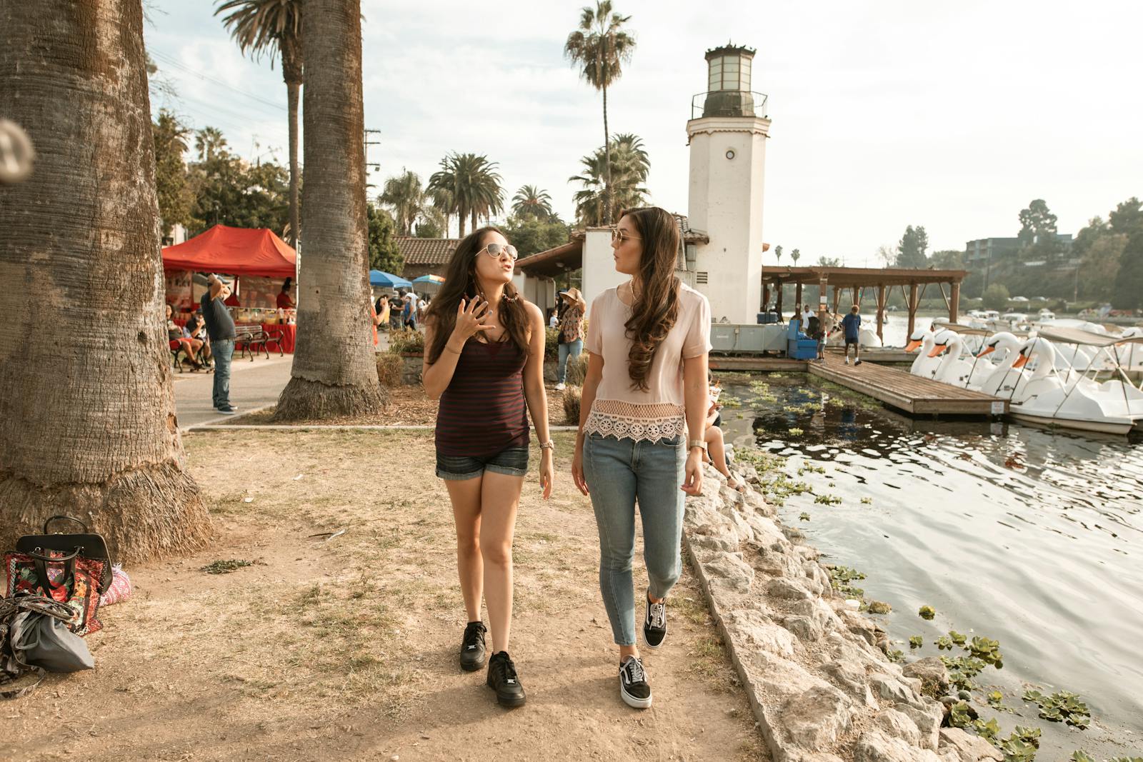 Two young women enjoy a lively conversation while walking by a lakeside lighthouse on a sunny day.