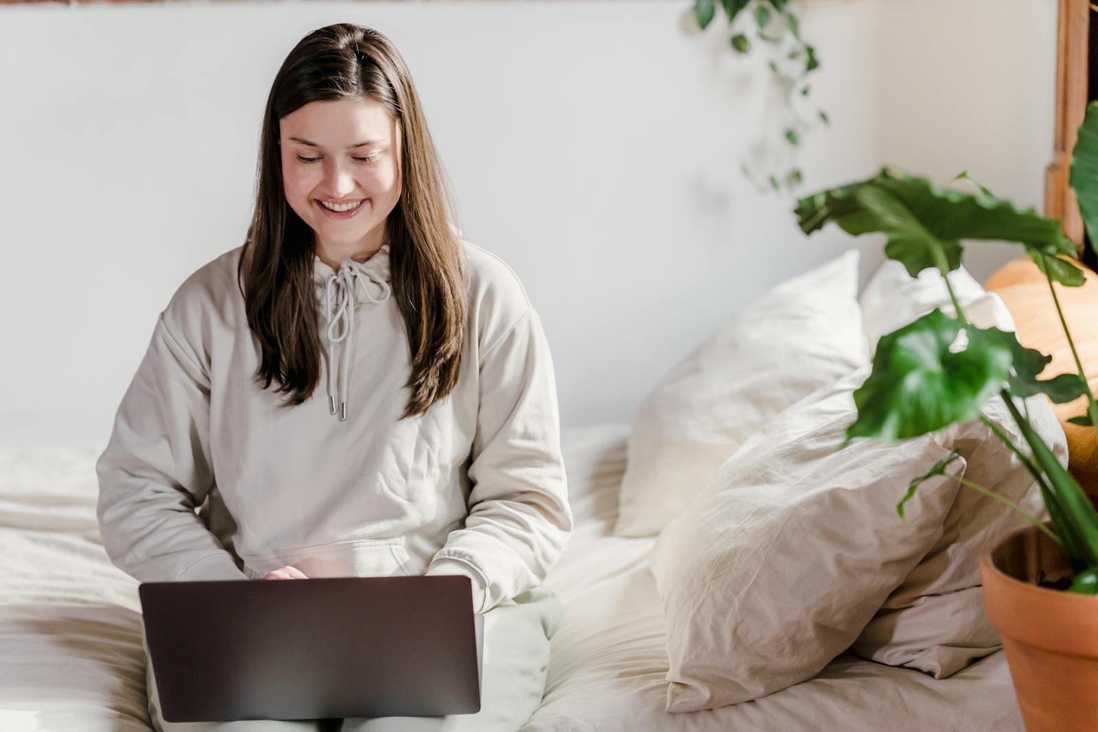 A cheerful woman using a laptop in a cozy bedroom with plants.