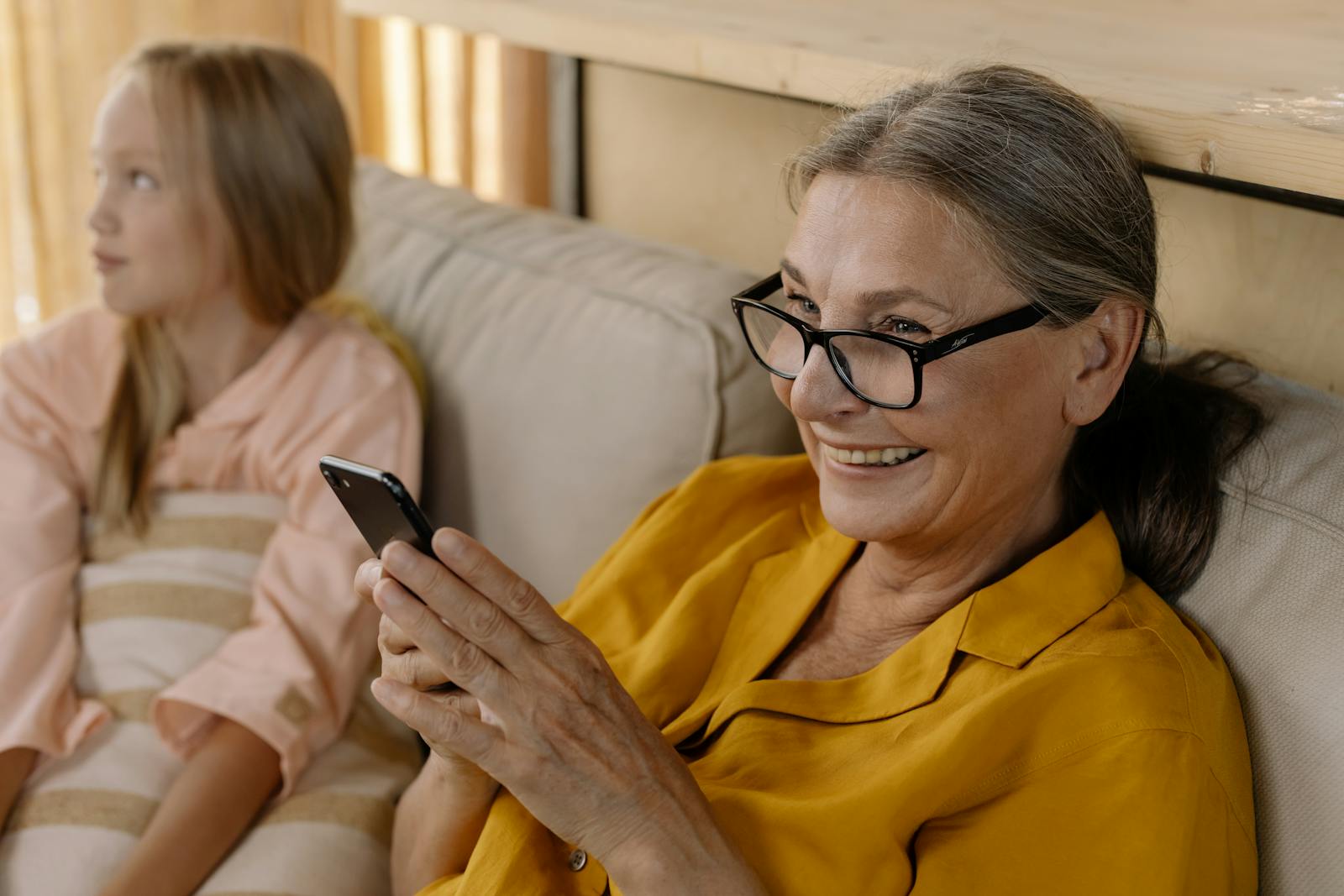 Senior woman in glasses smiling while using smartphone indoors, sitting with a child.