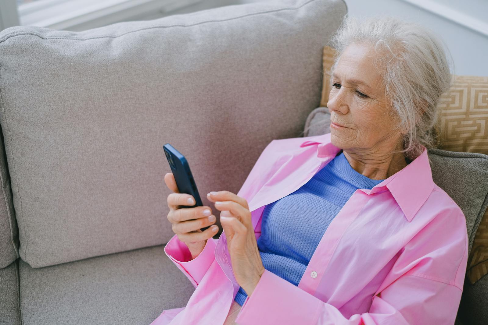 Elderly woman relaxing on sofa while using smartphone for communication.