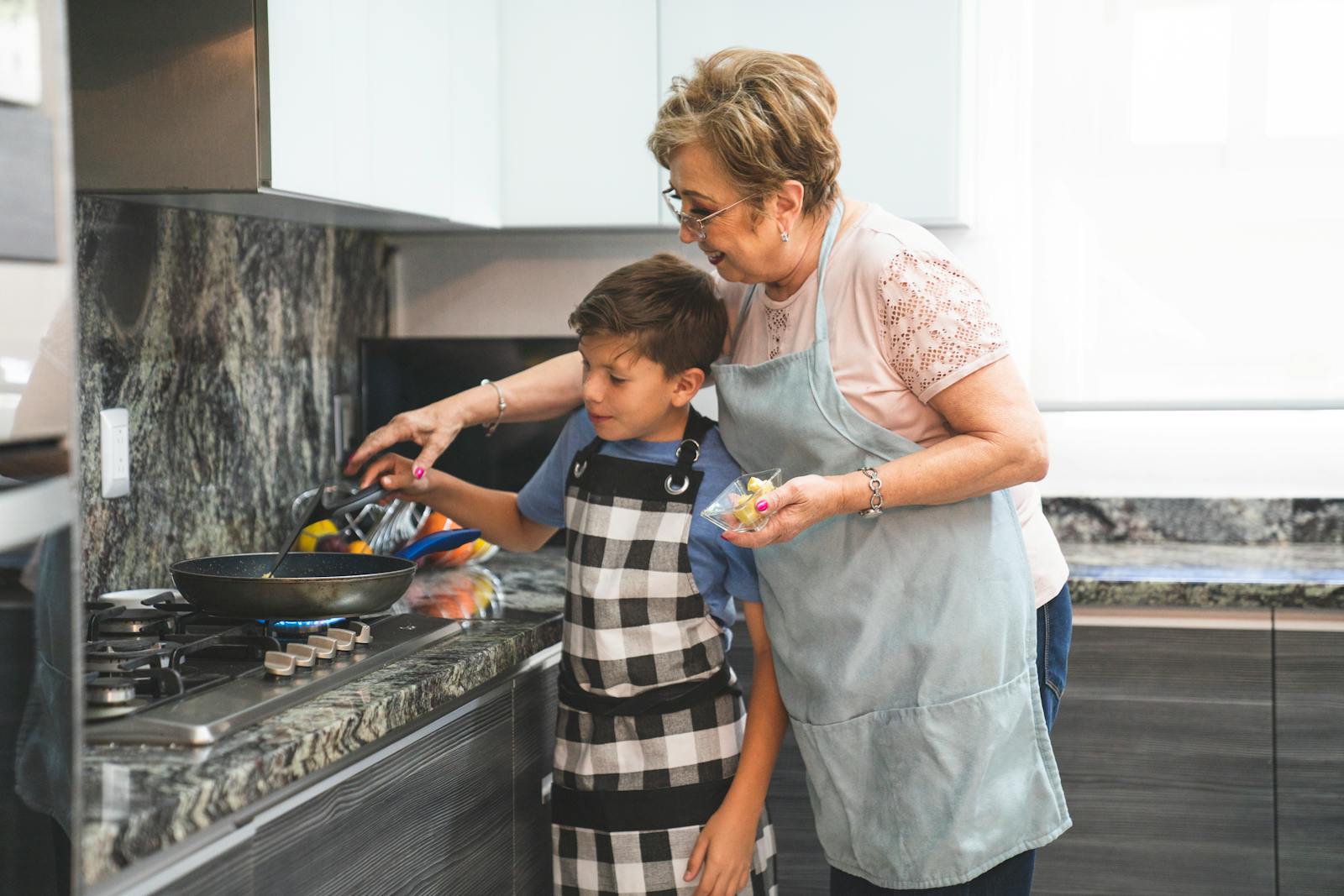 Lovely moment of a grandma teaching her grandson to cook. Both wearing aprons, standing by the stove.