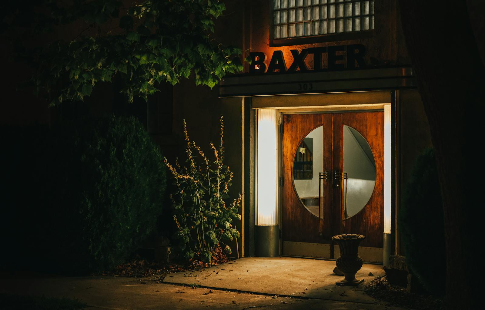 Illuminated wooden door entrance at night with greenery and potted plants outside a building.
