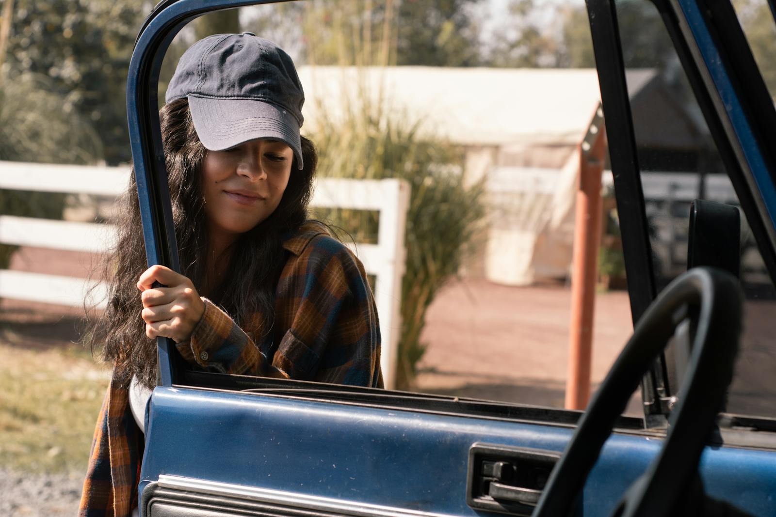 Casually dressed young woman in a cap opening a door of a blue car during the day.