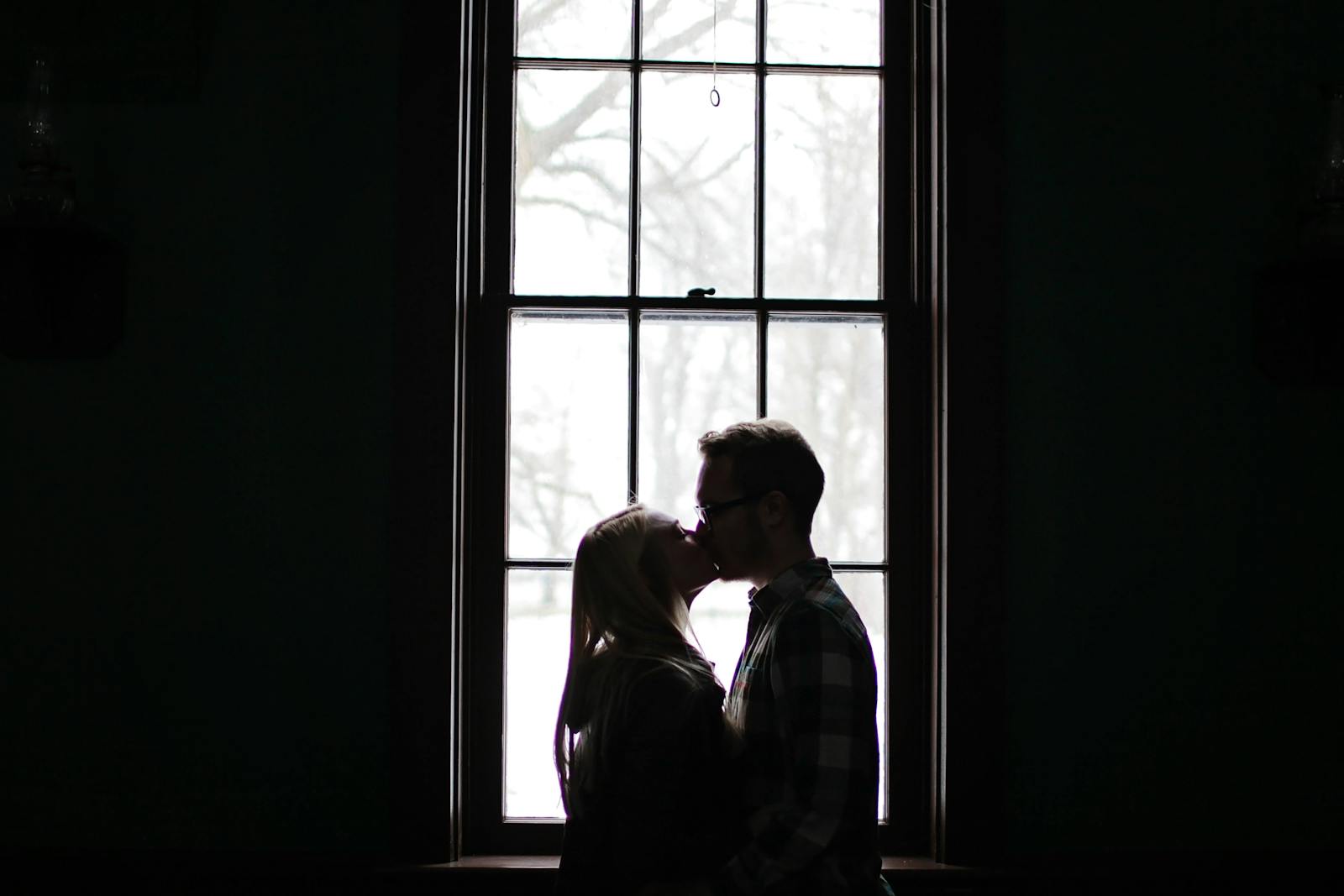 A romantic silhouette of a couple sharing a kiss in front of a large, backlit window.