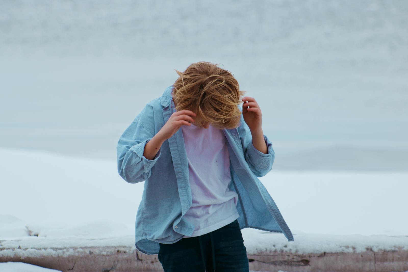 A fashionable boy in a blue shirt stands on a snowy beach, facing down.