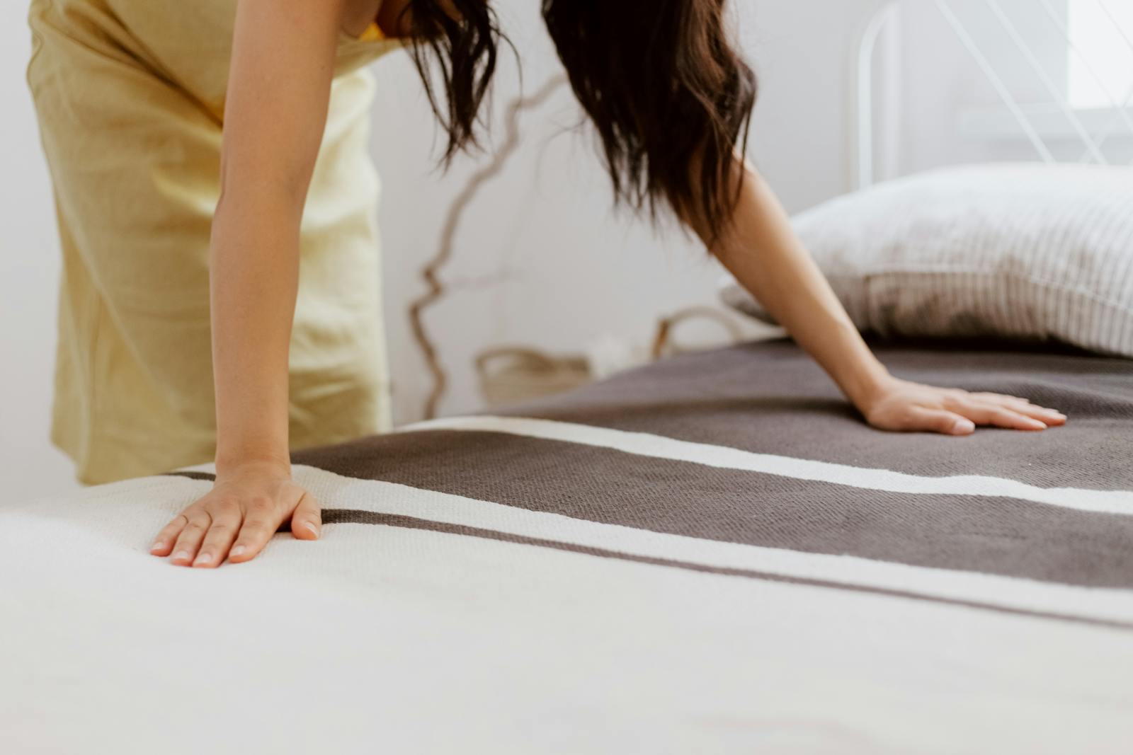 A woman making a bed adorned with striped linens in a bright, minimalist bedroom.