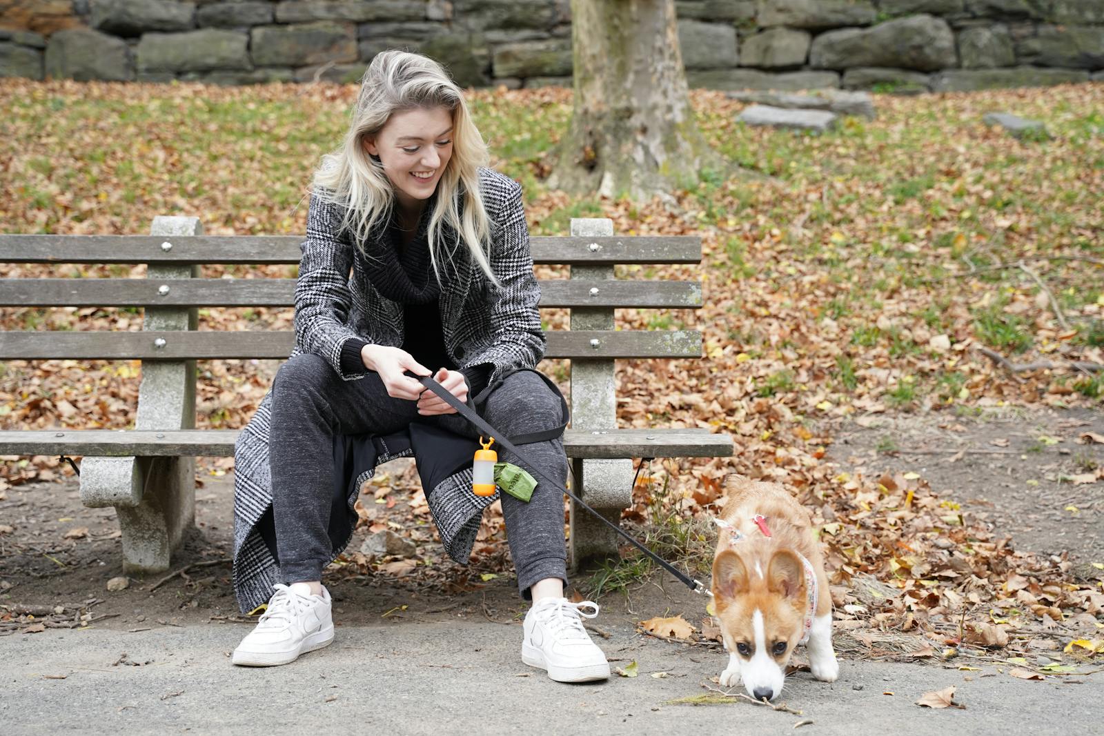 A woman sitting on a bench with her Welsh Corgi at Central Park in autumn, surrounded by fallen leaves.