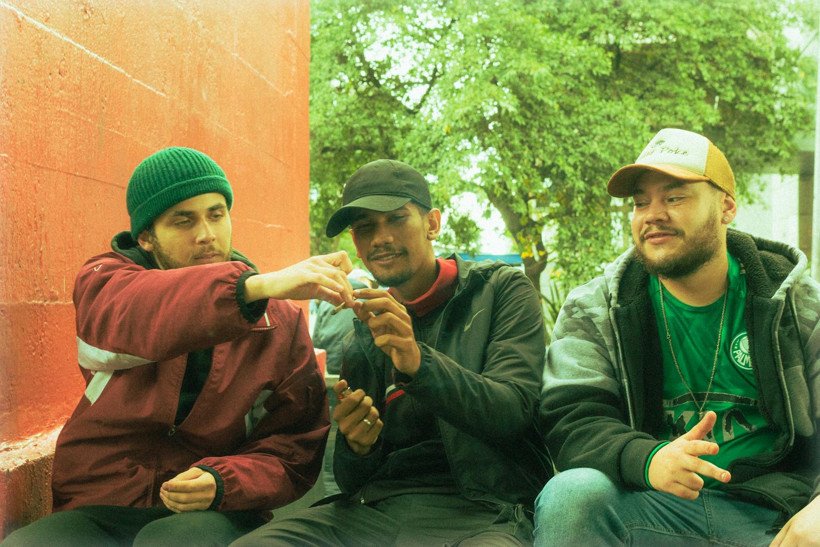 A group of three friends sitting by a red wall outdoors, sharing a cigarette and laughing together.