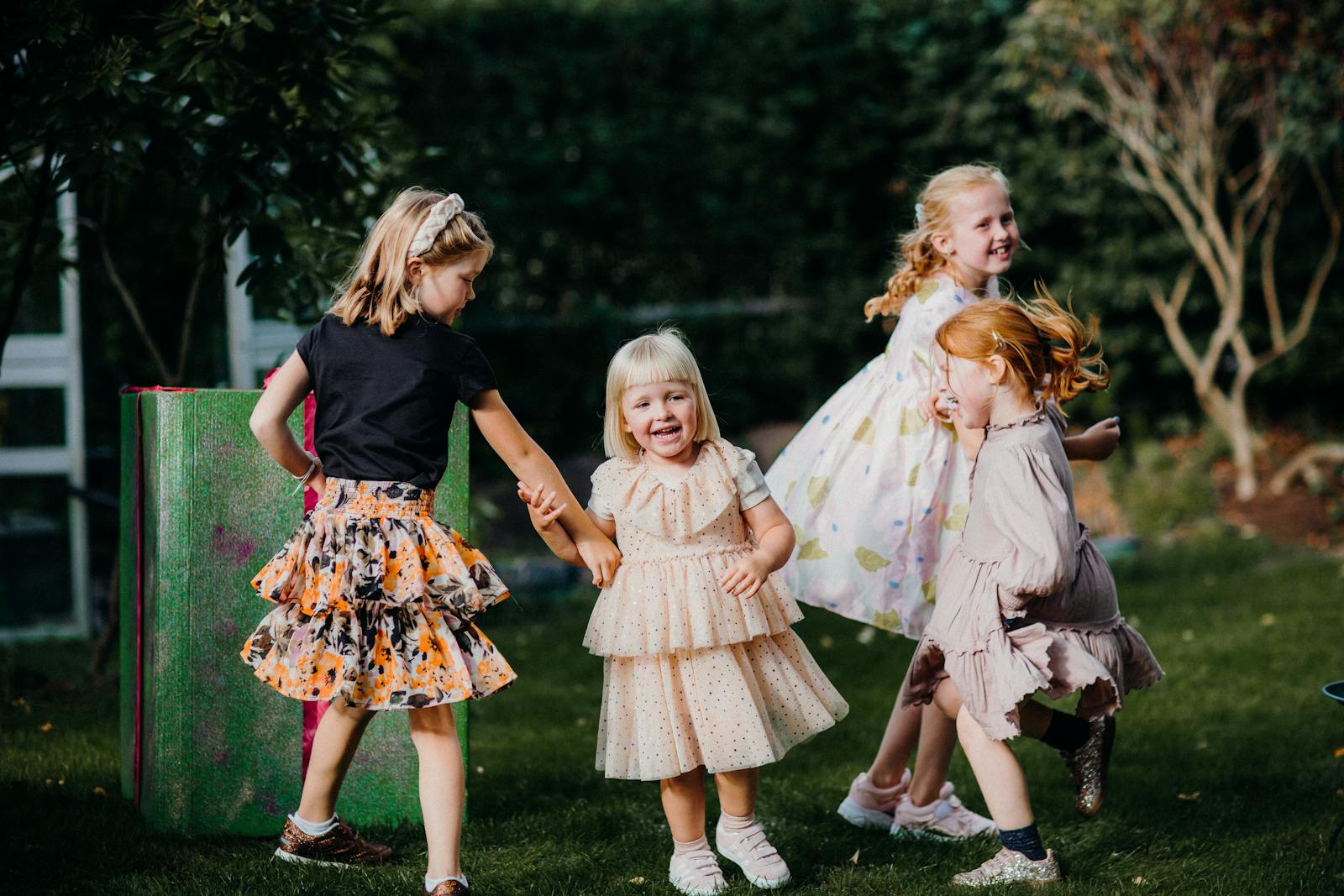 Four joyful children play in a garden, wearing colorful dresses and enjoying a sunny day.