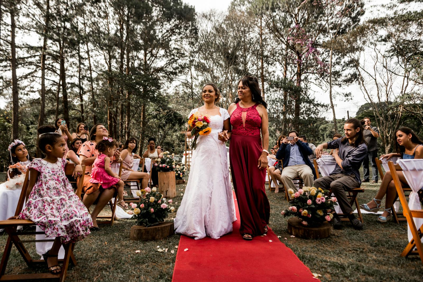 A joyful bride walking with her mother on a red carpet at an outdoor wedding ceremony surrounded by guests.