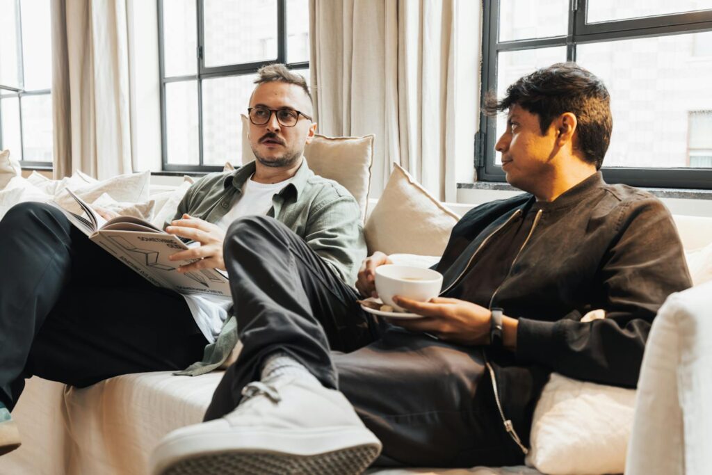 Two men sitting on a cozy sofa, one reading a magazine, enjoying coffee in a bright living room.