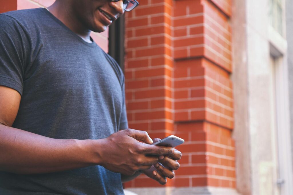 A smiling man checks his smartphone while leaning against a brick wall outdoors.