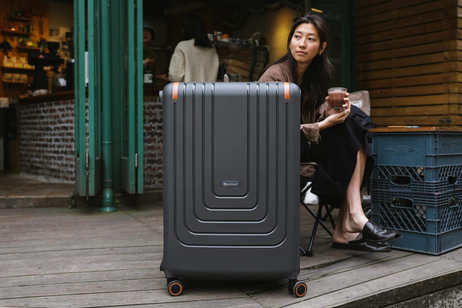 Asian woman at café holding coffee, seated with suitcase in urban setting.