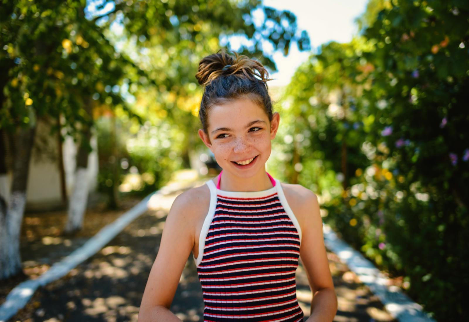 A cheerful teenage girl smiling in a sunny garden walkway with lush greenery.