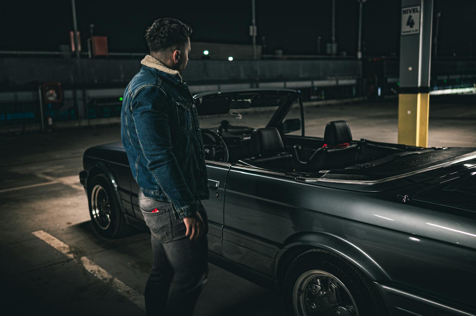 A man in denim jacket stands beside a convertible in a dimly lit parking garage at night.