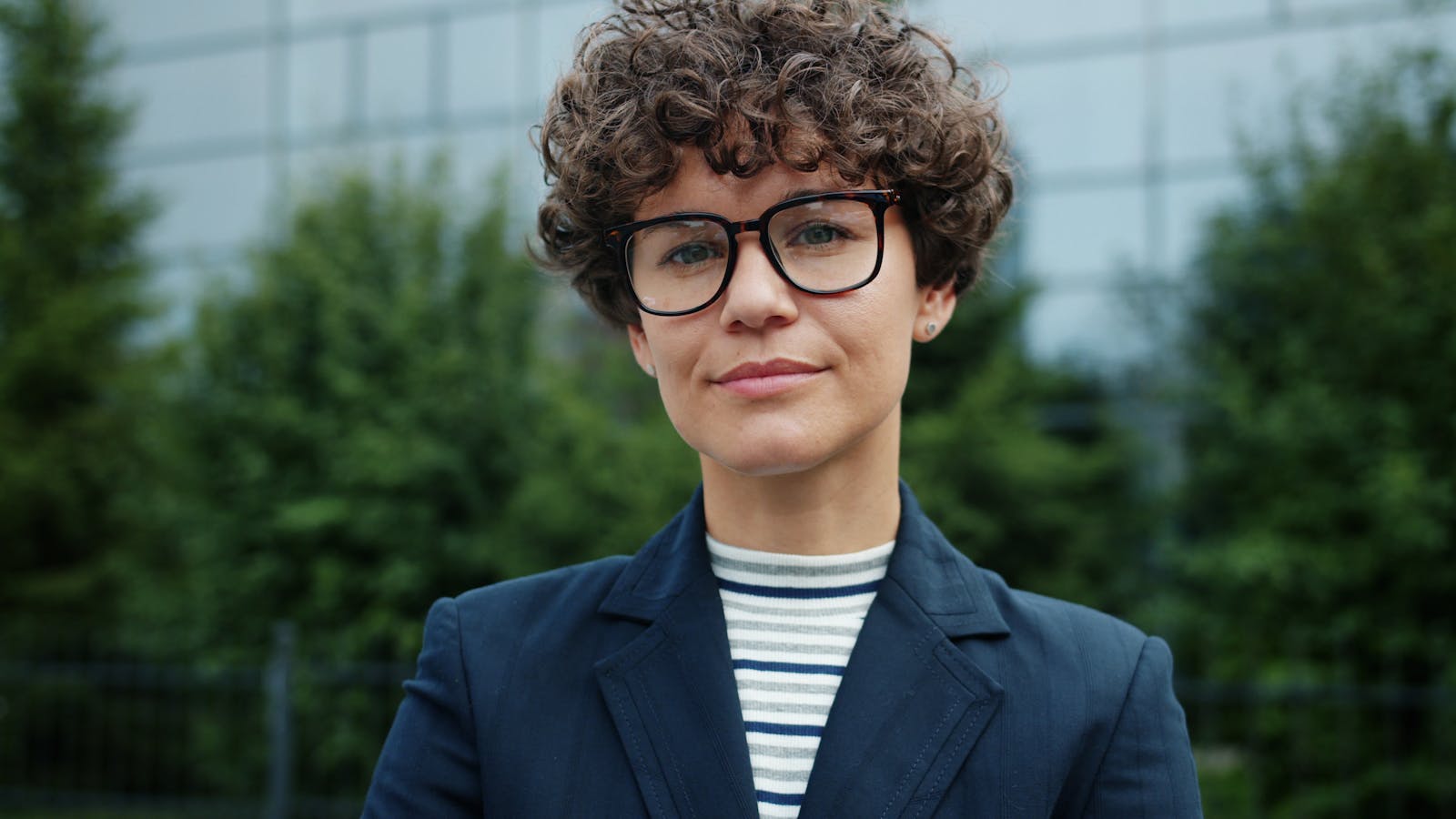 Portrait of a confident woman in business attire with curly hair and glasses outdoors.