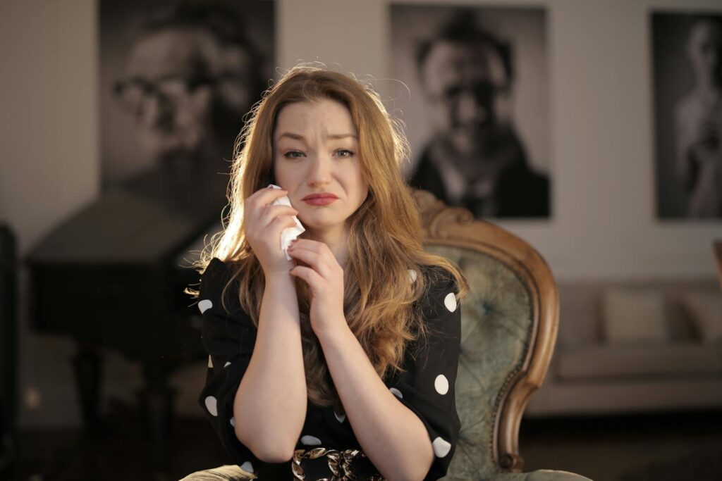 A woman sitting in a vintage chair indoors, showing emotions and holding tissues.
