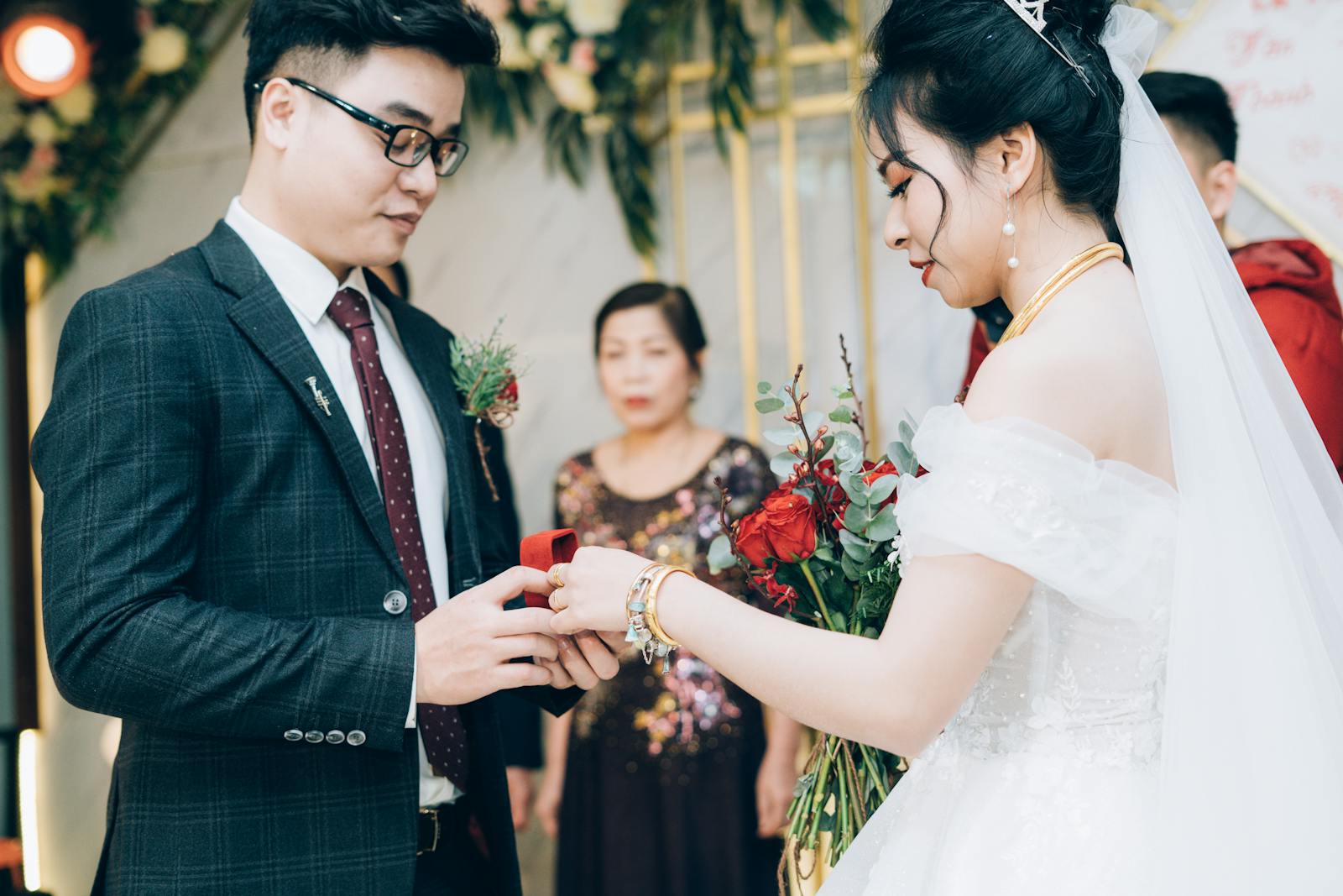 Side view of crop ethnic bride with makeup in dress with bridal bouquet and groom in suit with eyeglasses preparing rings during marriage ceremony in decorated hall behind parent