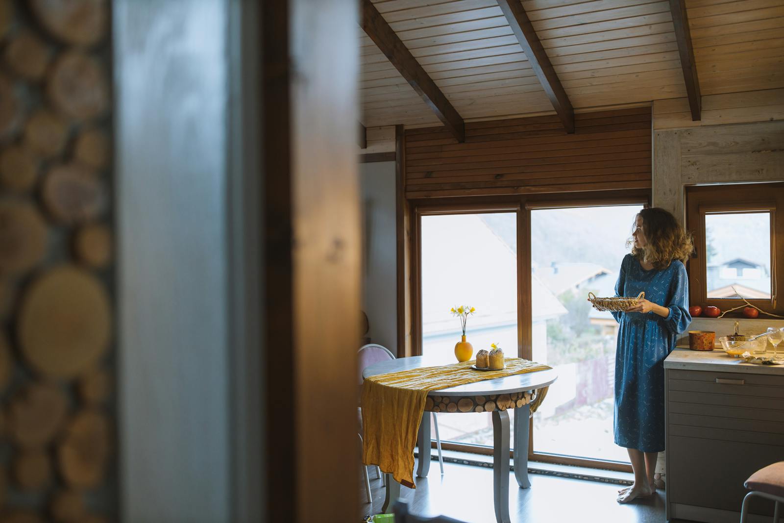 A woman in a blue dress stands indoors holding a wicker basket near a sunlit window.