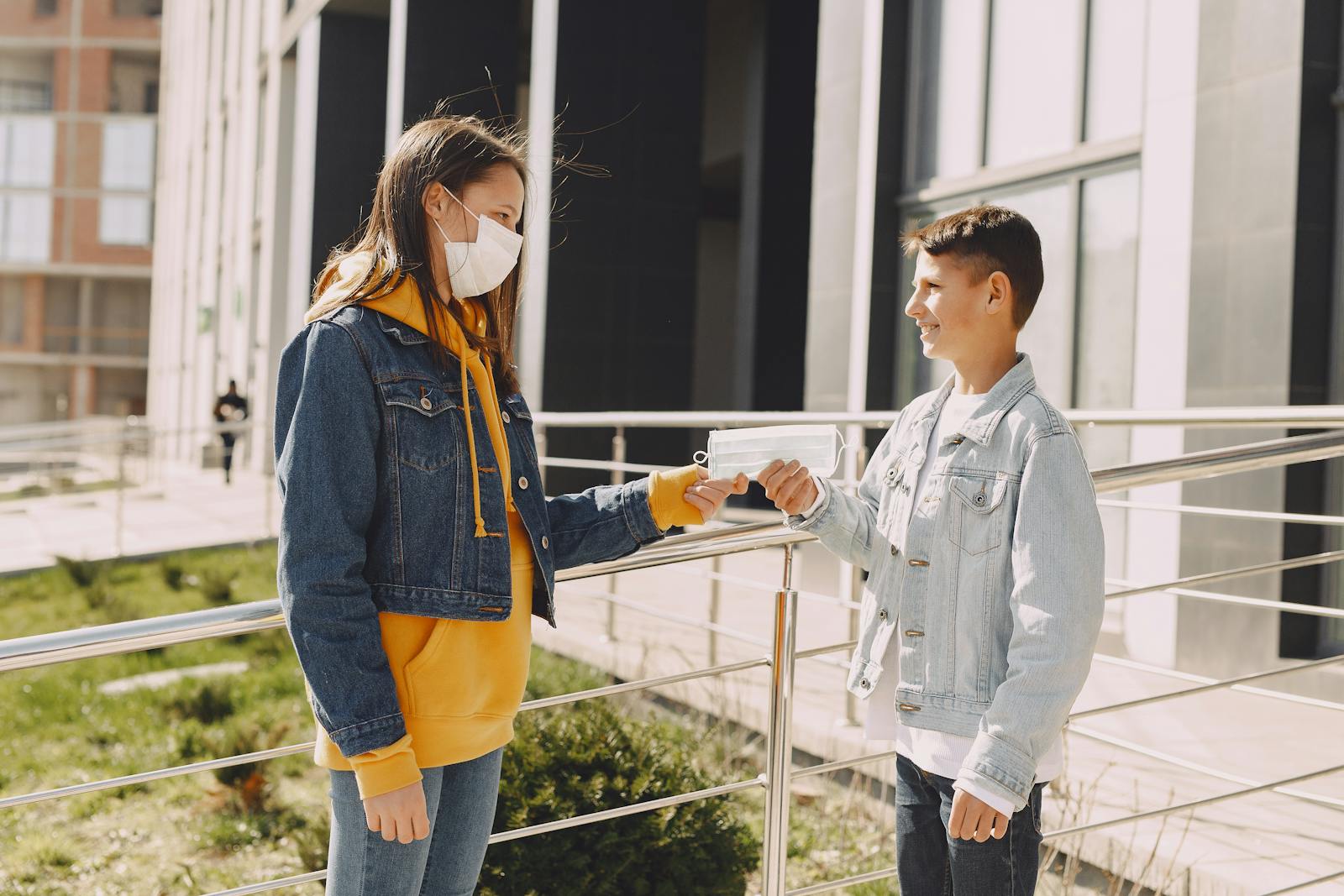 Side view of happy young boy in casual clothes smiling and standing against elder sister in medical mask on city street while taking face mask