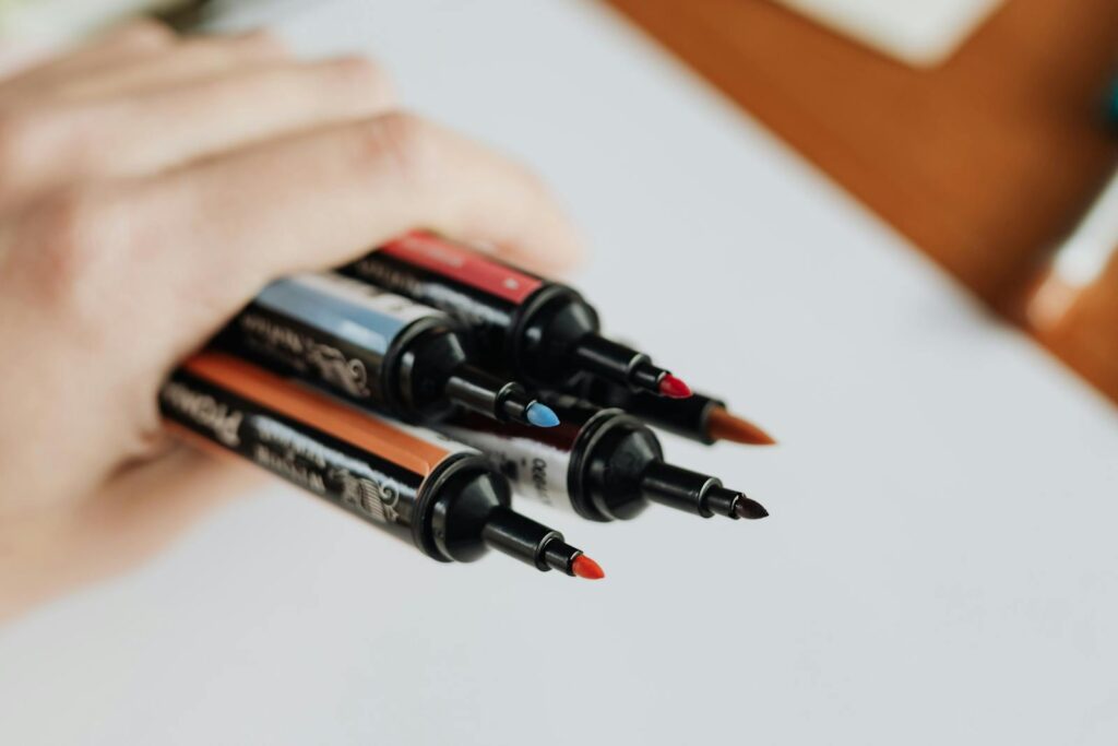 Close-up of a hand holding four assorted color markers against a soft focus background.