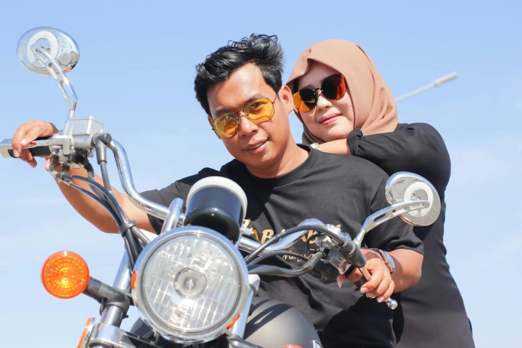 Smiling couple enjoying a ride on a classic motorcycle under clear blue skies, wearing sunglasses and a hijab.