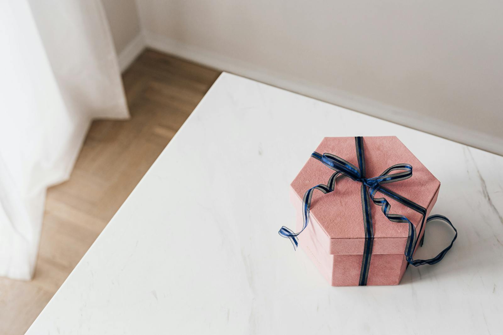 From above of packed pink hexagonal gift box tied with blue ribbon placed on marble table against beige wall and wooden floor and white curtain