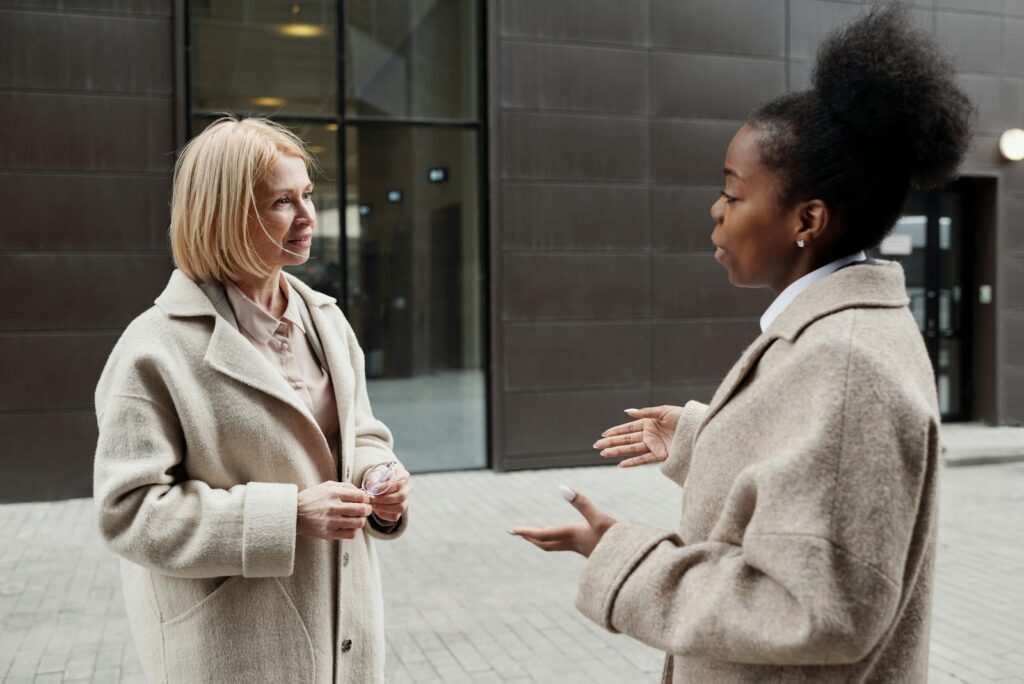 Two businesswomen in coats converse outside a modern office building.