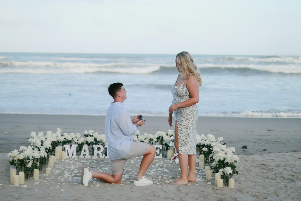Man proposes to woman on beach with flowers.