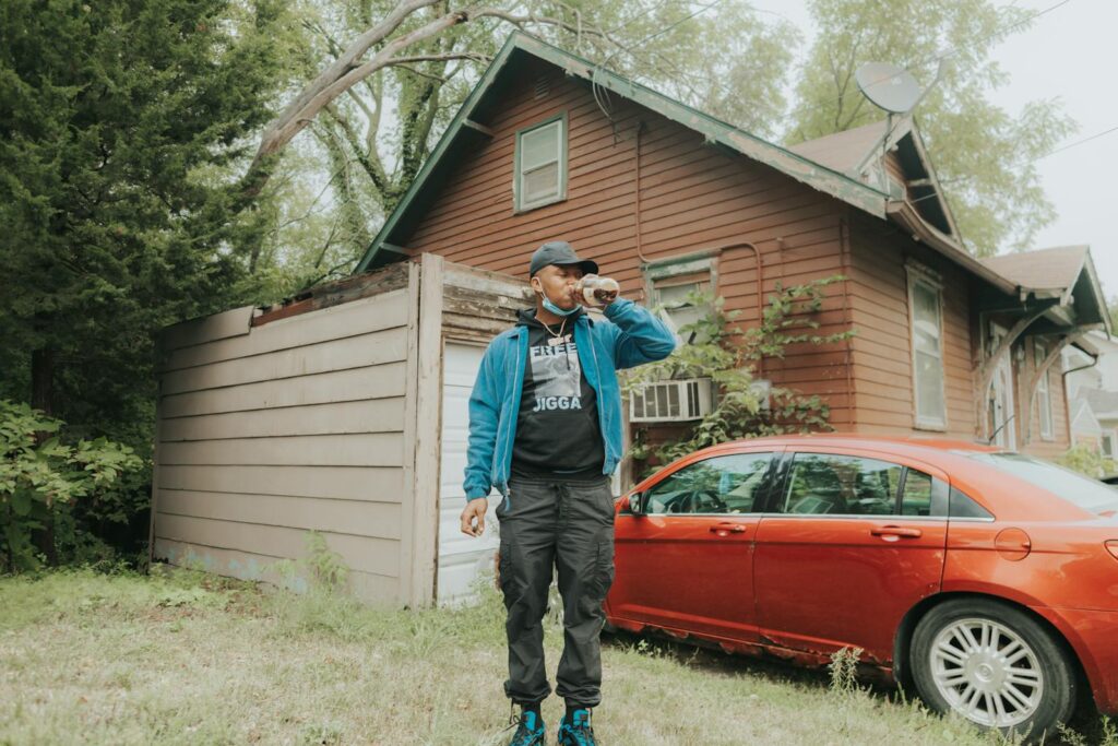 Man in blue jacket drinking outside next to a red car and house.