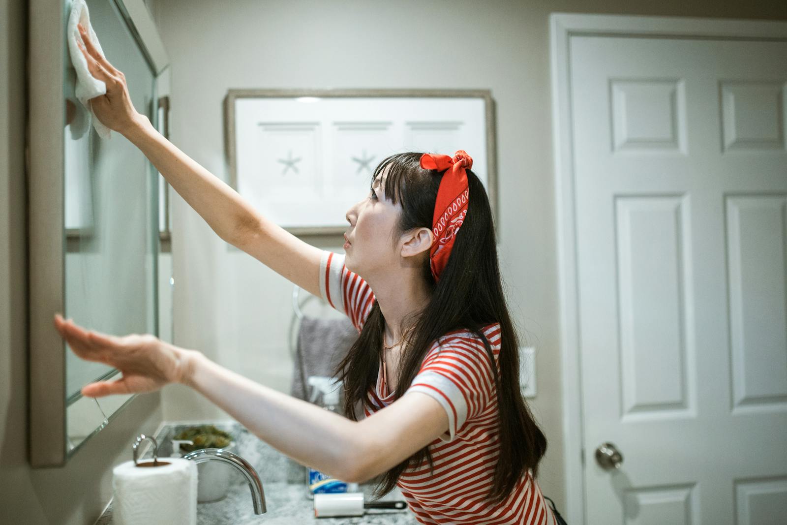 Woman in a striped shirt cleaning a mirror in a bathroom with a red headband.