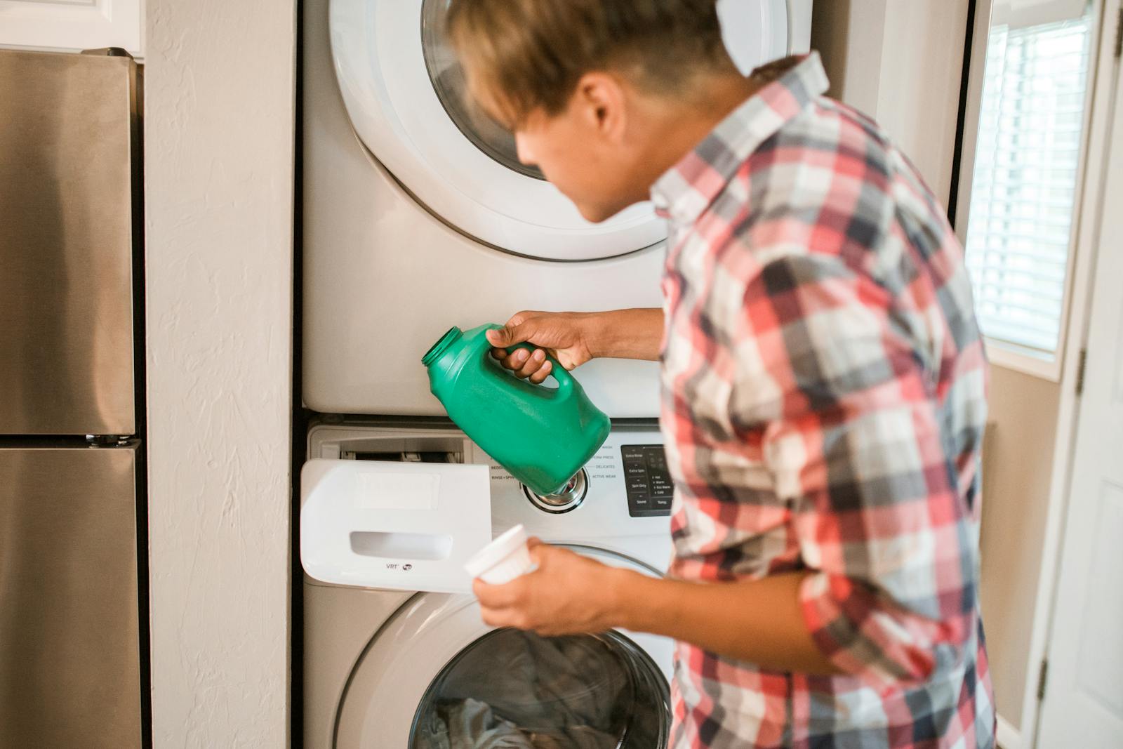 A man in a plaid shirt pours detergent into a front-loading washing machine, engaging in a household chore.
