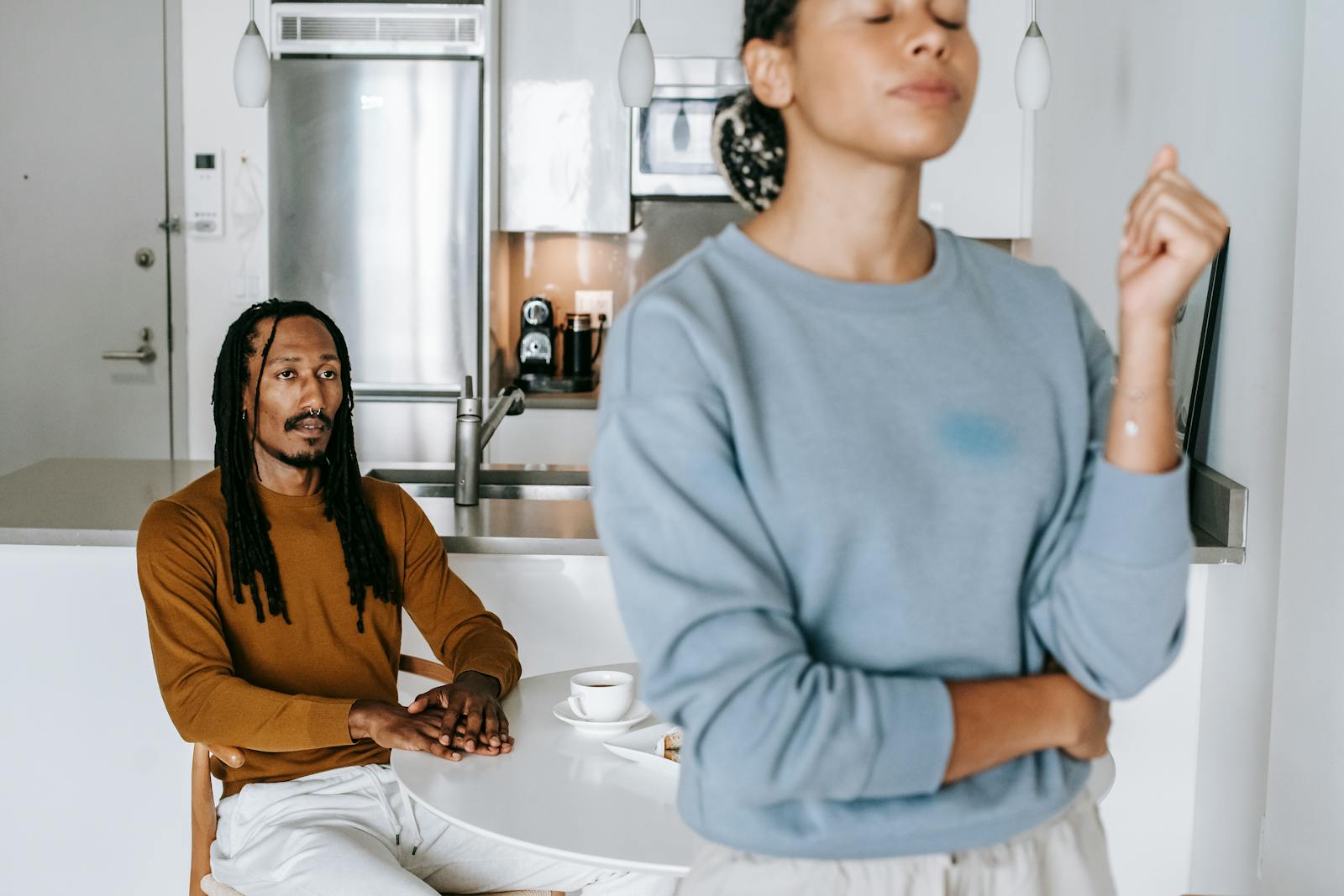 Young black man sitting at table while having conflict with standing near table woman in light kitchen