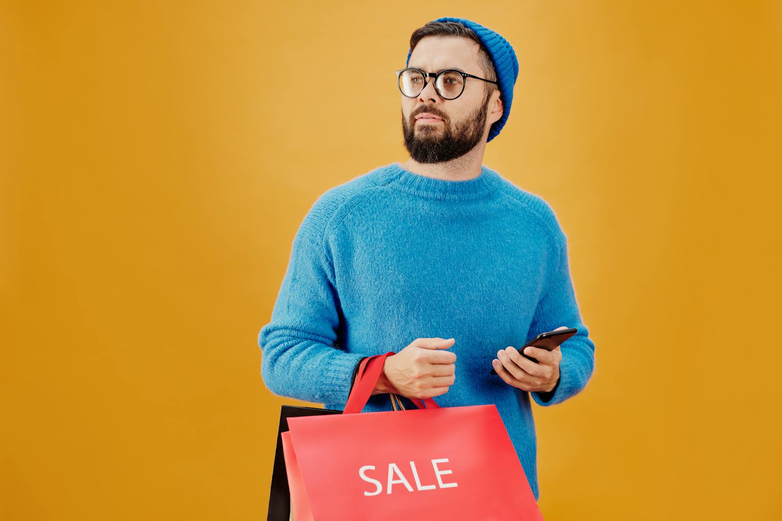 Stylish man in blue holding sale bags and smartphone, looking away.