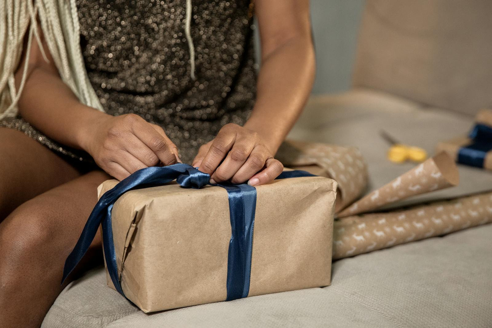 Close-up of a woman wrapping a gift box with blue ribbon on a couch.