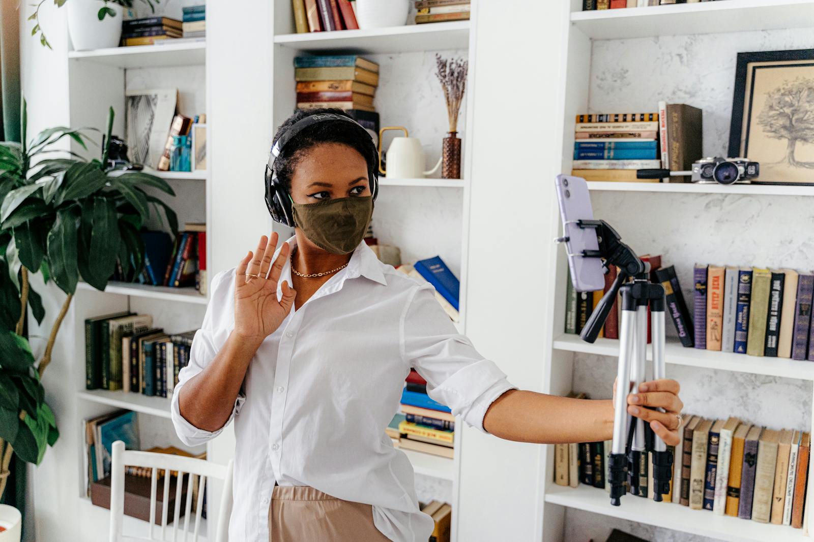 Woman wearing mask waves at smartphone on tripod in home library. Streaming or video call setup.