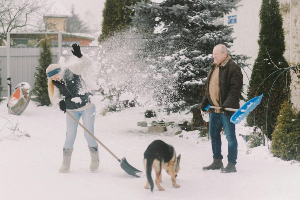An elderly couple having fun while shoveling snow in a snowy backyard with their dog.