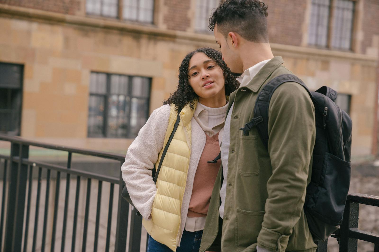 A young couple stands together with backpacks in a city setting, discussing warmly.