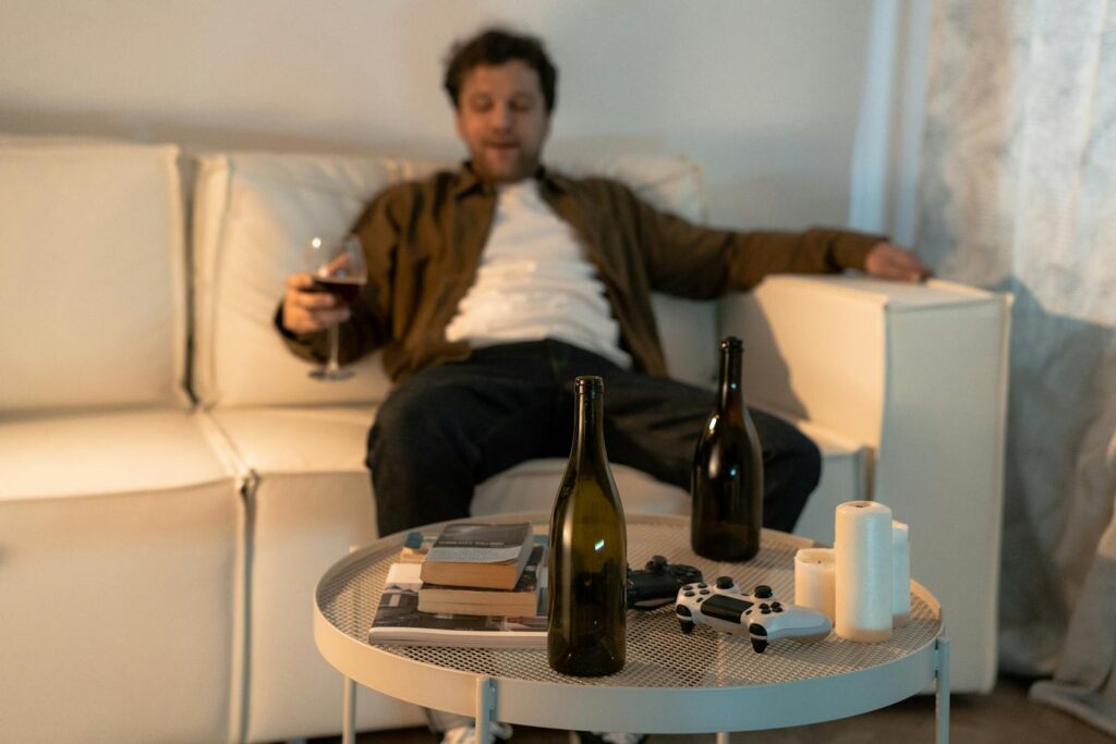 A man sits on a couch with wine bottles, a game controller, and books, expressing relaxation or contemplation.