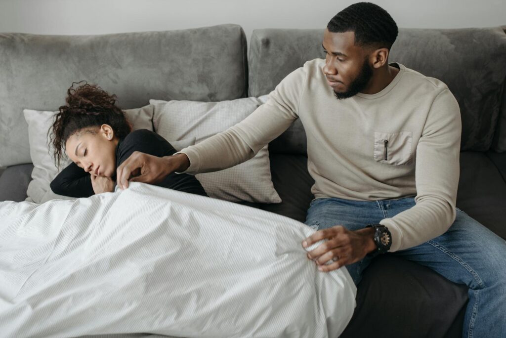 A concerned man tends to his unwell partner resting on the sofa. Emotional scene of support and care.
