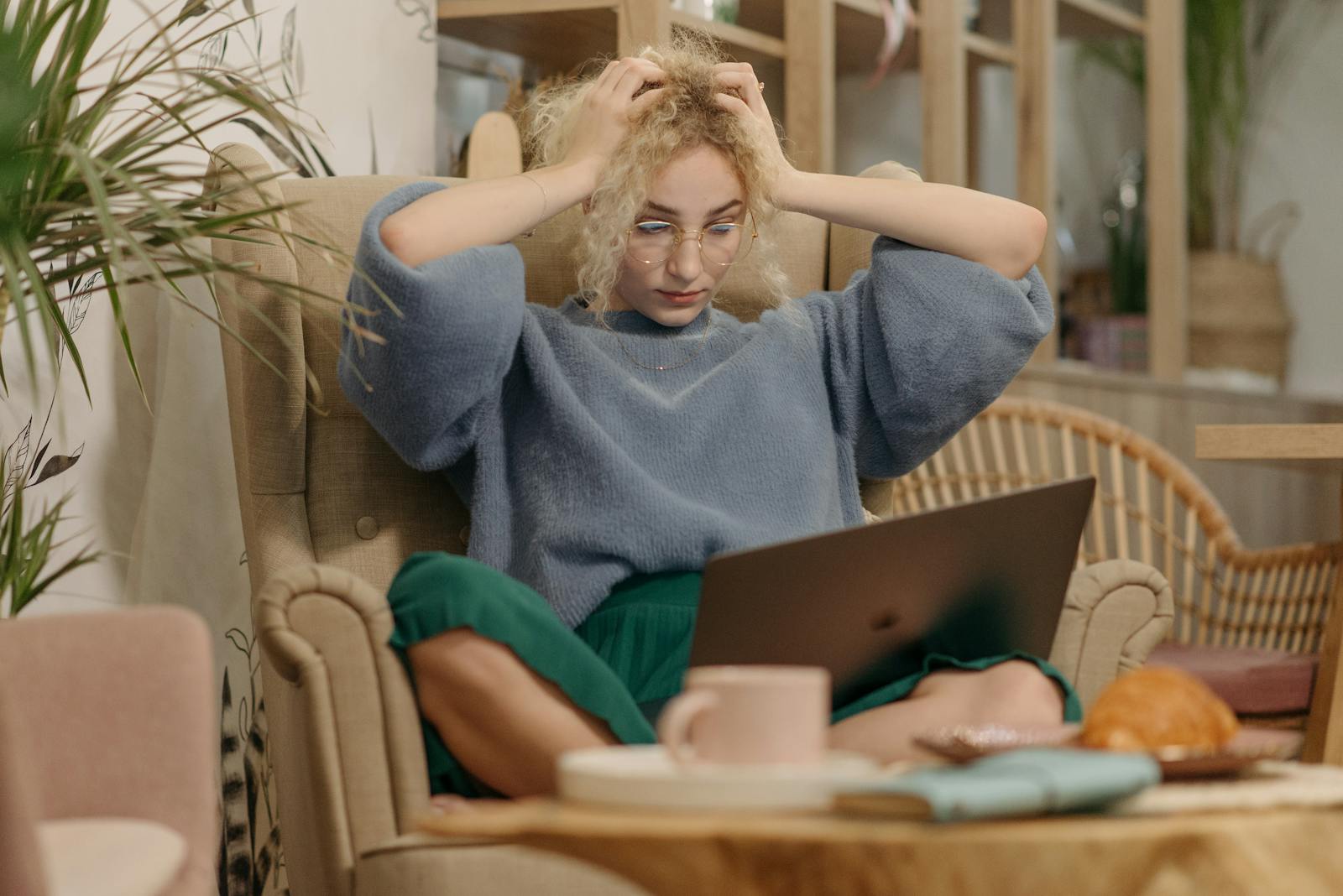 A young woman looking stressed while using her laptop indoors, sitting in an armchair with hands on her head.