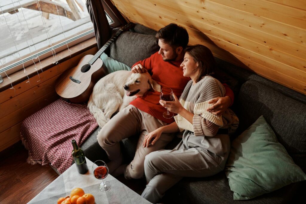 A couple enjoys wine and cuddles with their dog in a cozy cabin setting.