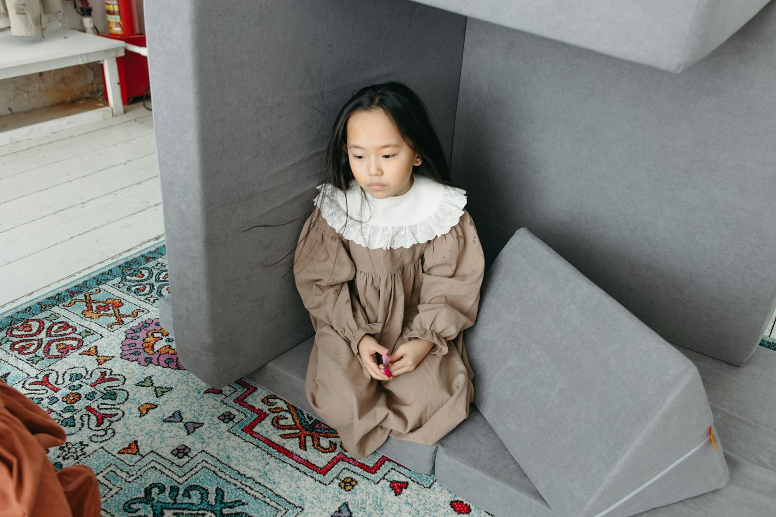 Child in a vintage dress sitting indoors on a colorful carpet in a cozy room.