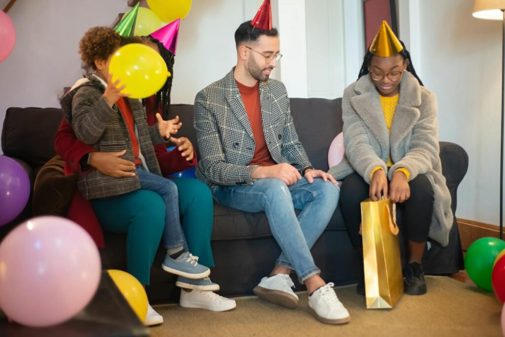 A happy family celebrating a birthday indoors with balloons and gift bags, wearing party hats.