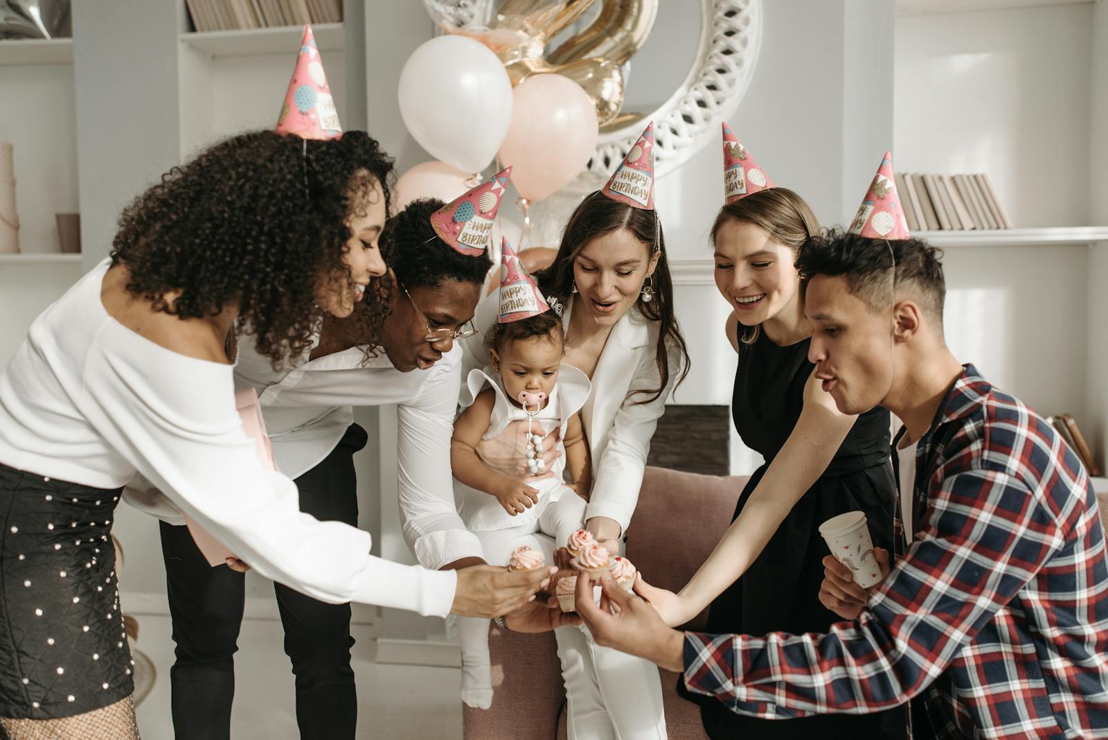 A diverse group celebrating a child's birthday indoors with cupcakes and party hats.