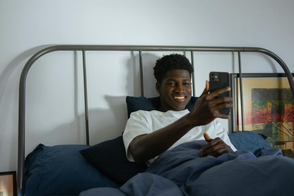 A man smiling while using a smartphone in a modern bedroom setting, illustrating relaxation and connectivity.