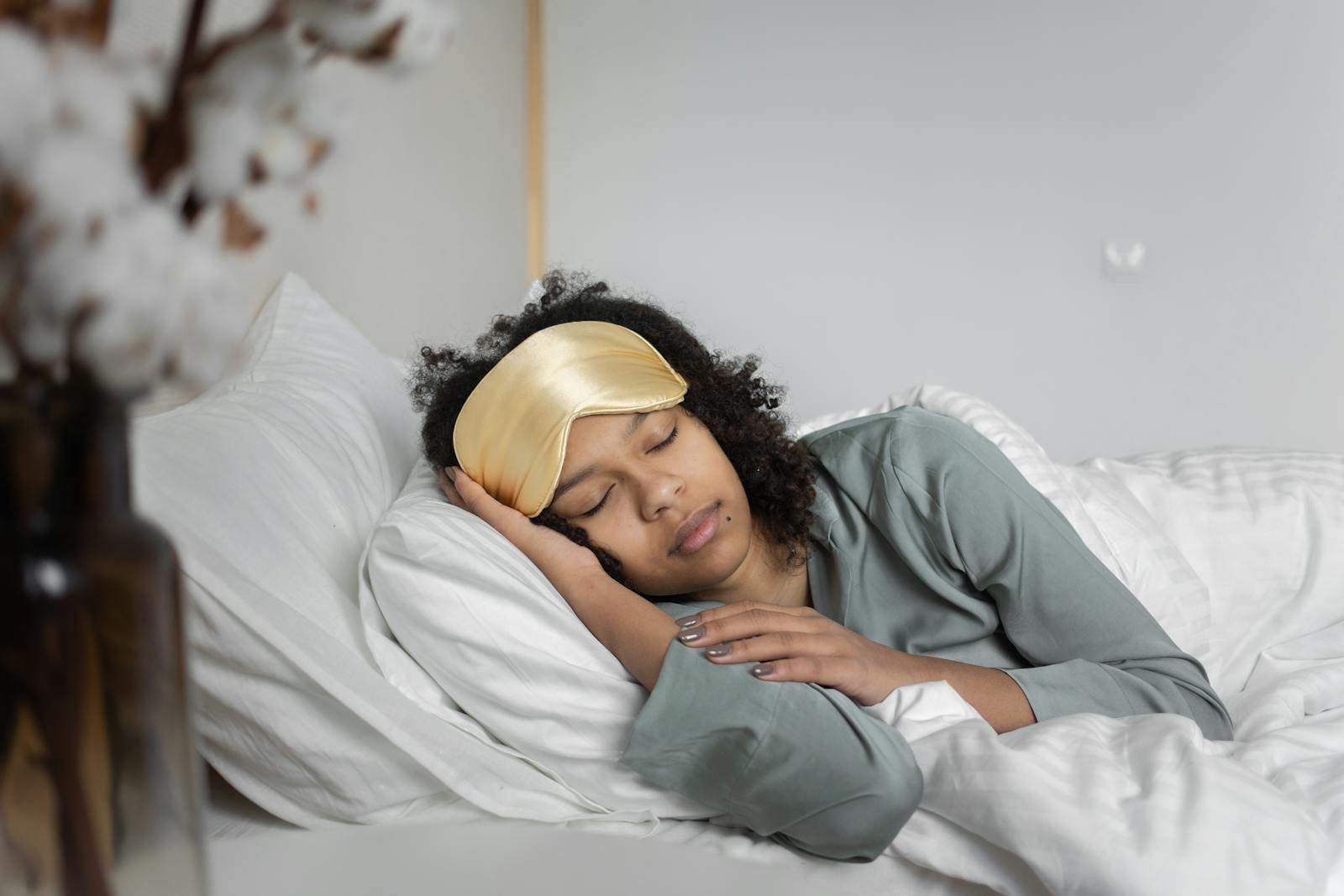 A young woman peacefully sleeps with a sleep mask, nestled in cozy white bedding.