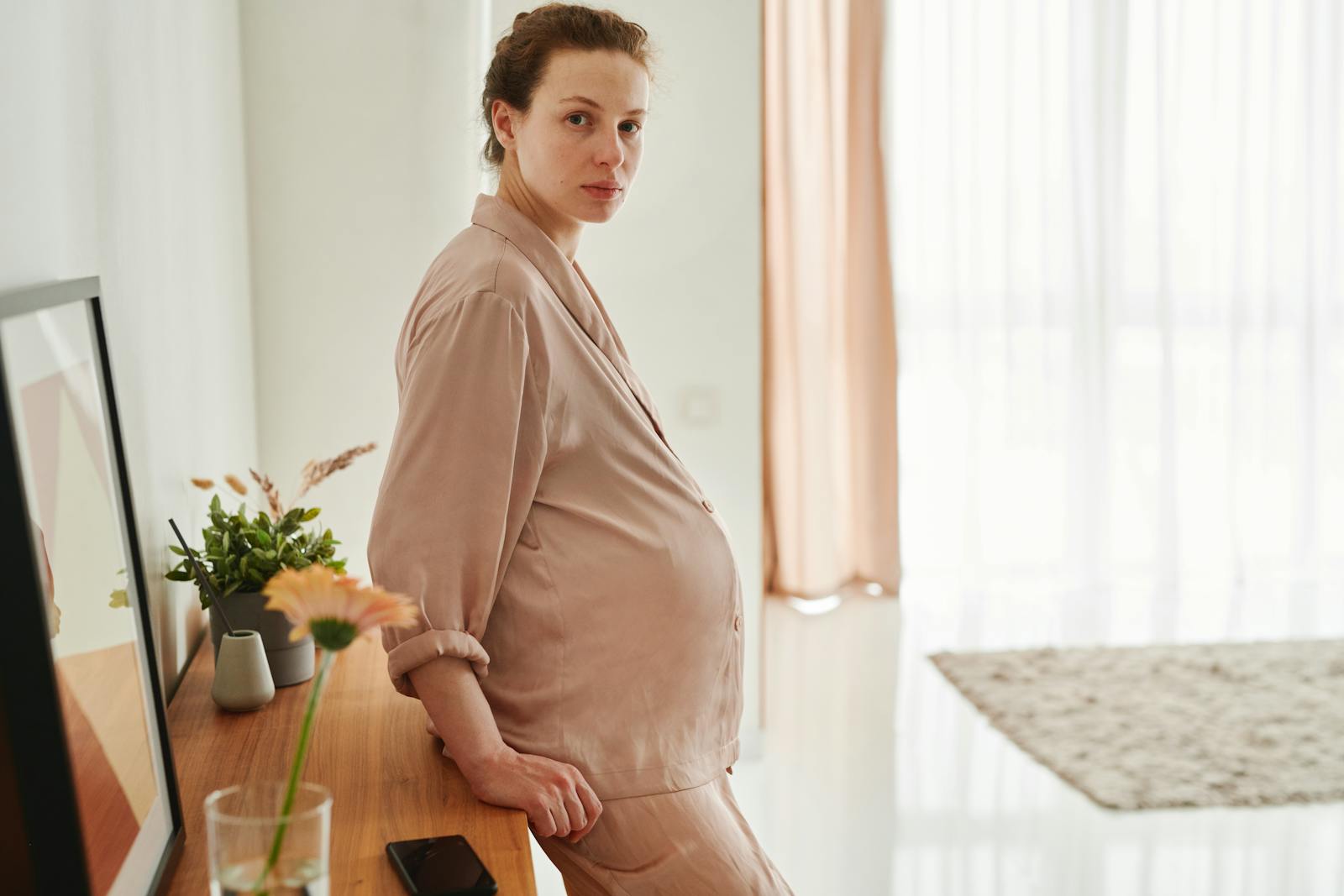 A pregnant woman wearing sleepwear leans on a wooden drawer in a bright, serene room.
