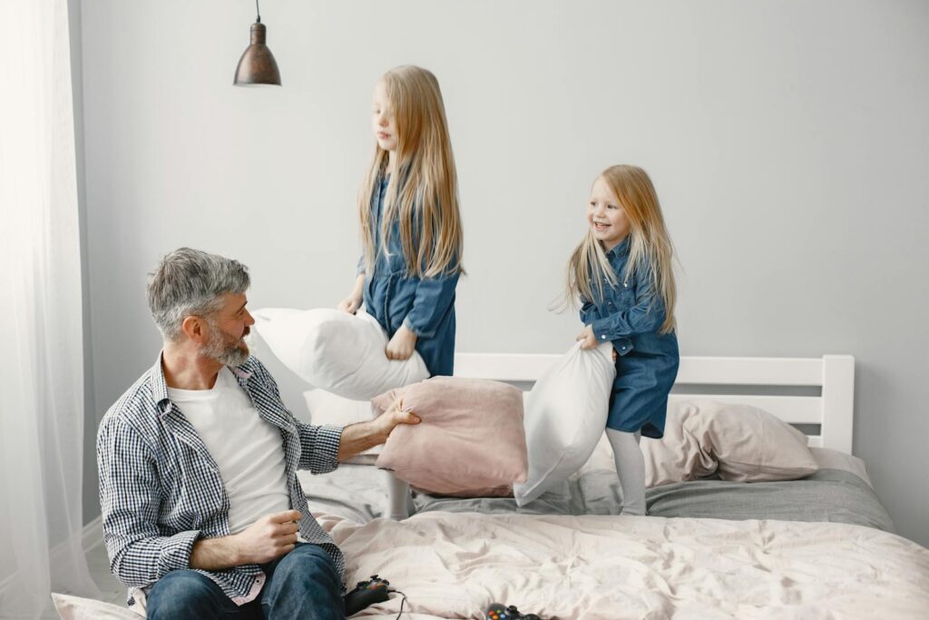 Grandfather and granddaughters having fun pillow fight on bed indoors.