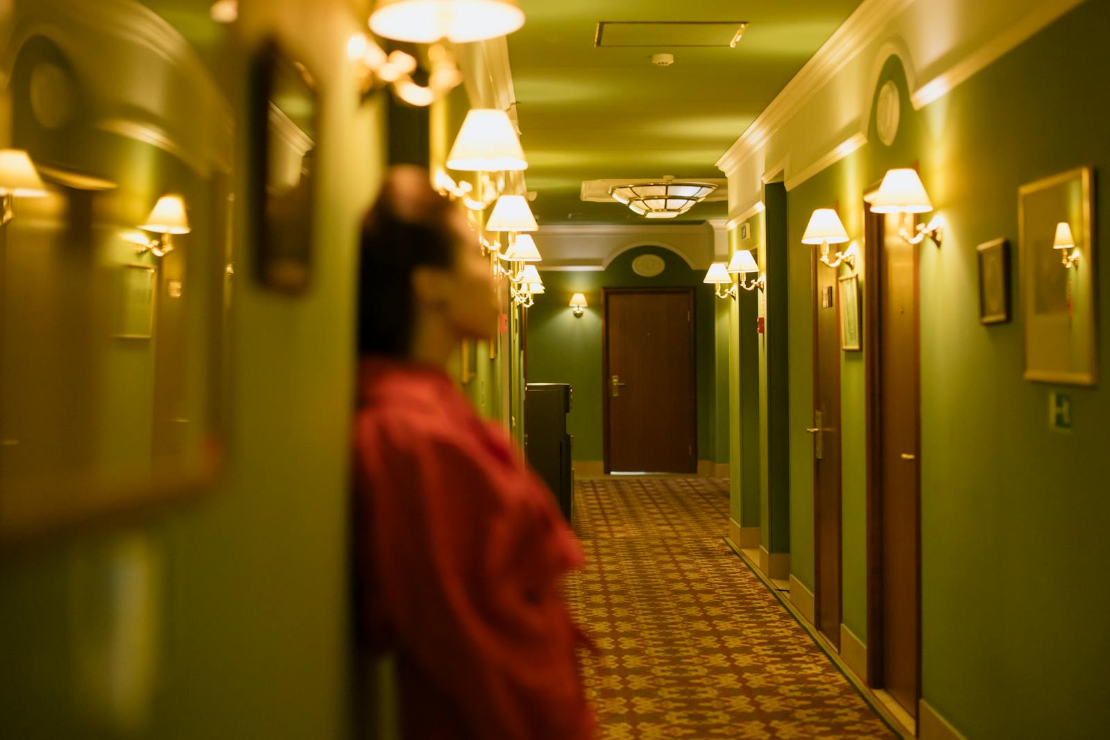A woman in a red dress leans against the wall in a dimly lit hotel hallway.